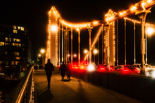 Pedestrians walking on the beautifully illuminated Chelsea Bridge at night. There is also a lot of vehicular traffic on the bridge, and the headlights of cars glow in the misty night.