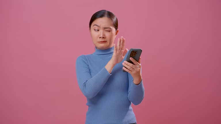 Asian woman feel scared looking at smartphone and covering eyes by hands standing isolated over pink background.