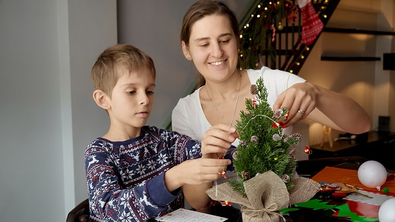 Happy little boy with mother decorating small Christmas tree with garlands and lights. Winter holidays, family time together, kids with parents celebrating