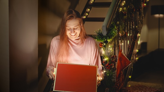 Portrait of excited smiling woman looking inside of glowing shiny gift box with Christmas present.