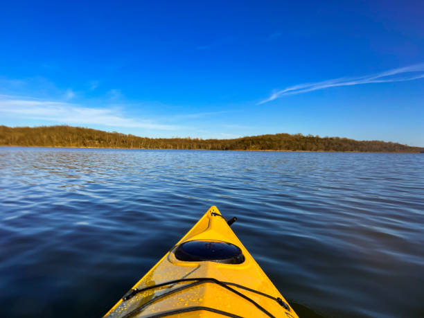 The photo captures the serene and relaxing atmosphere of a summer day on the lake, where a yellow kayak floats gently on the clear water. The kayak provides a bright contrast to the natural colors of the surroundings, inviting the viewer to imagine paddling and exploring the lake. The horizon is lined with green trees, creating a sense of depth and harmony. The sky is a deep blue, dotted with white clouds that reflect the sun’s rays. This is a photo that shows the beauty and tranquility of nature, and the joy of outdoor recreation.