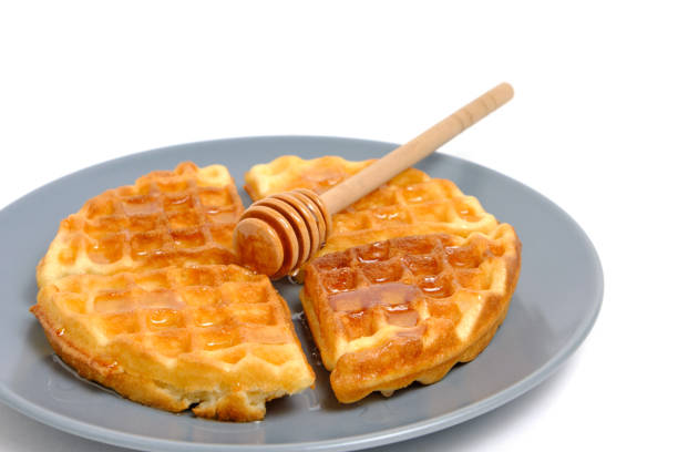 Soft muffin waffles poured with fresh honey using honey spoon, in plate on white background