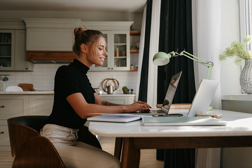 Side view of attractive young woman sitting at the desk and using laptop while working at home