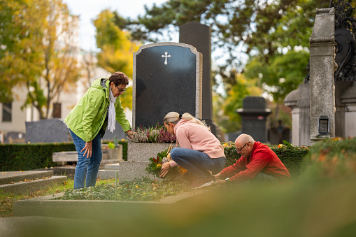 senior couple, parents and mature adult daughter, preparing family grave with all saints decoration on autumn day at old graveyard in vienna