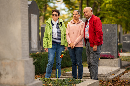three people, mature adult daughter and senior parents, standing together at family grave of old graveyard on sunny day in autumn