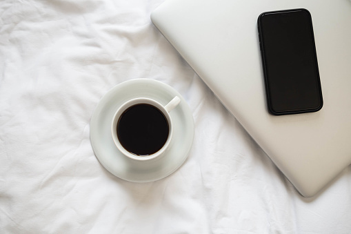 Cup of black coffee, laptop and smartphone on unmade white bed, work from home concept.