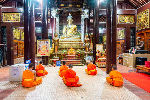 Chiang Mai,Thailand-March 20 2023: Illuminated by lights inside the ancient teak wood structure,monks chant during evening prayers,before the golden Buddha,within Chiangmai's Old City walls.