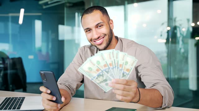Satisfied businessman at workplace happy with financial achievement results, holding phone and dollar bill money in hand