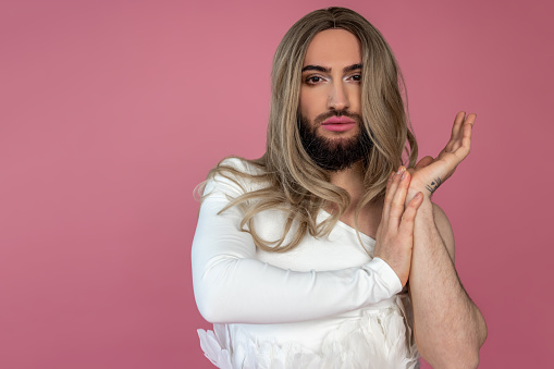 Attractive handsome transgender model wearing blonde wig and white dress looking at camera with confident expression posing isolated over pink background