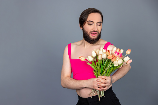 Attractive romantic brunette transsexual wearing pink top holding bouquet of spring flowers admires beauty posing isolated over gray background with copy space