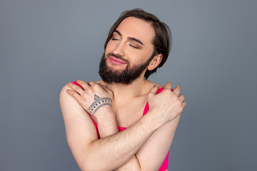Satisfied self loving brunette transsexual wearing pink top hugging himself with smile and pleasure posing isolated over gray background