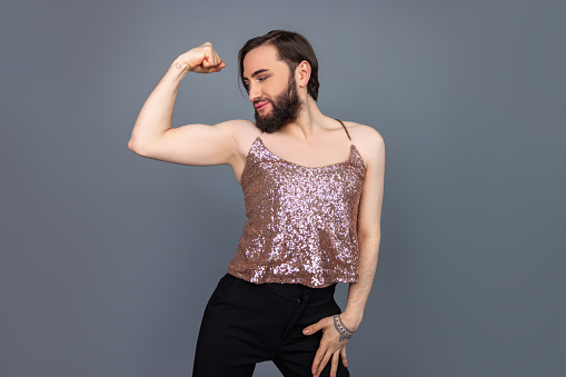 Slim attractive transsexual person wearing shiny tank top having perfect body raised arms showing biceps posing isolated over gray background