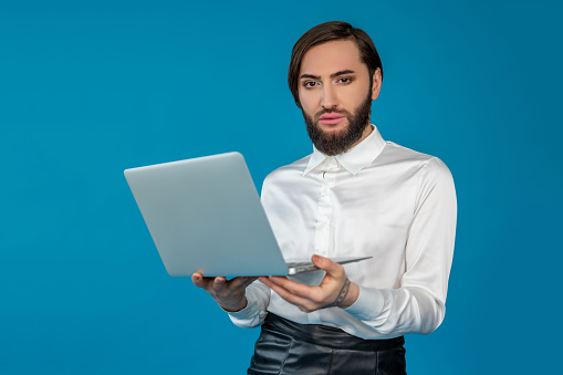 Self confident transgender man wearing white shirt working on laptop browsing internet looking at camera posing isolated over blue background