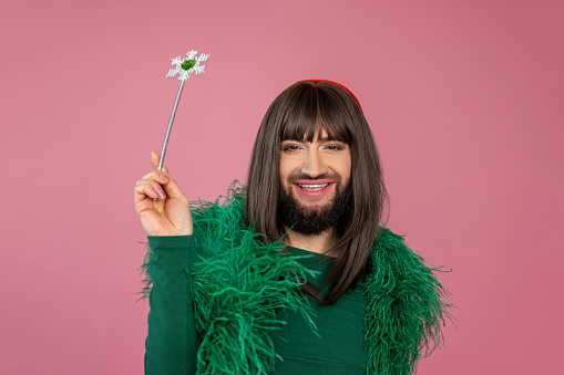 Cheerful smiling transgender with makeup and wig wearing green feather party dress playing with magic wand posing isolated over pink background