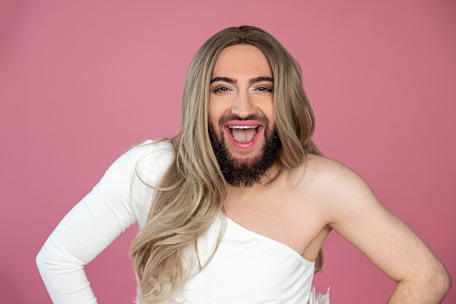 Extremely happy transgender wearing blonde wig and white dress keeps hands on hips laughing out loud posing isolated over pink background