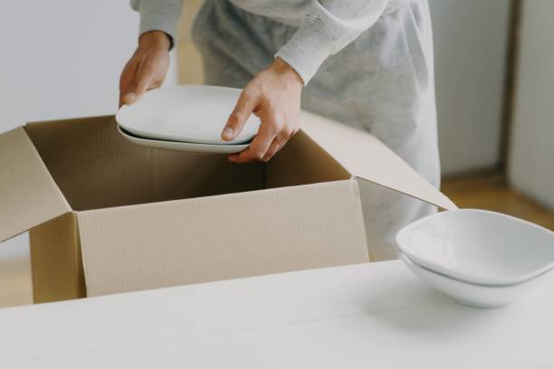 istock Hands carefully unpacking white dishes from a cardboard box on a table in a bright room. 1802528275
