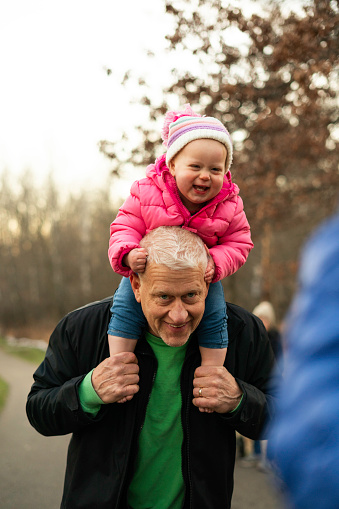 Grandpa makes a silly face into a camera while his barefoot  granddaughter laughs sitting on his shoulders, Minnesota