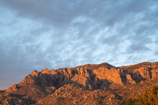 The Sandia Mountains illuminated by the light of the setting sun, casting the rocks in orange and pink colors, with a light sky with high, thin clouds above.