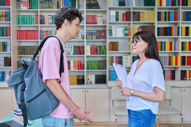 istock Middle-aged female teacher talking to male college student inside library 1802573589
