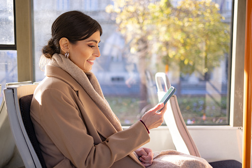 Beautiful young woman sitting on a bus, using a mobile phone while commuting to work. Happy young businesswoman talking on smartphone while traveling by public transport.