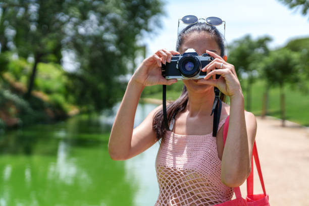 Female photographer in pink mesh top using a camera outdoors with greenery and lake in background.