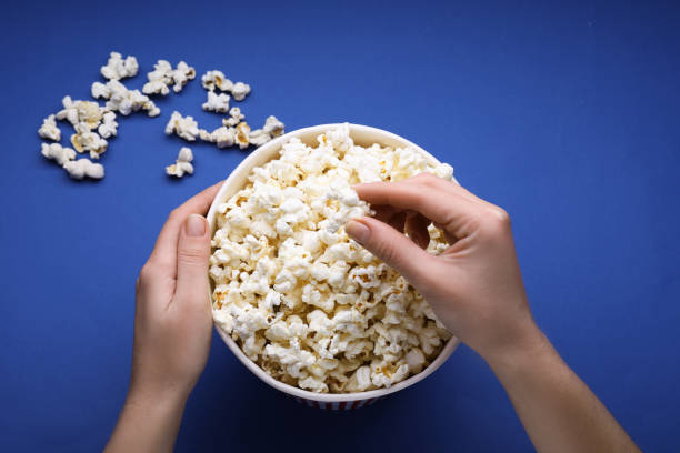 Woman taking fresh popcorn from bucket on blue background, above view