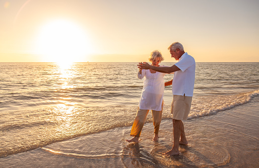 Happy senior man and woman old retired couple walking dancing and holding hands on a beach at sunset
