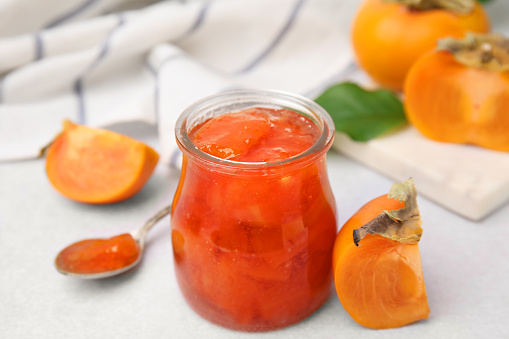 Jar of tasty persimmon jam and ingredients on white table