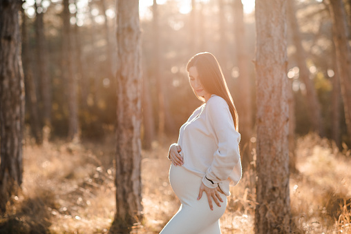 Cute smiling pregnant woman 25-29 year old wear knitted sweater holding her belly in woods outdoor. Maternity lifestyle. Autumn season.