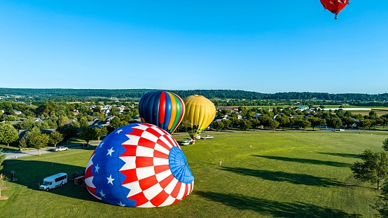 An Aerial View of 3 Hot Air Balloons Setting Up to Launch With One in the Air, on a Field in a Community, on a Sunny Summer Afternoon