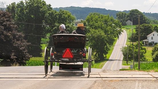 Ronks, United States – July 23, 2023: Ronks, Pennsylvania, July 23, 2023 - A Rear View of an Amish Couple in an Open Horse and Buggy, Crossing RR Tracks Heading Down a Rural Road