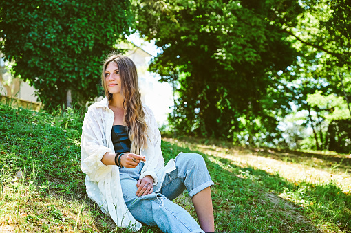 Young fashionable woman in a casual clothing sitting on the ground in a parkland.