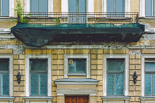 An old balcony in disrepair covered with a green professional construction protective net. Facade of a residential building in St. Petersburg, Russia.