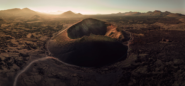 El Cuervo Volcano Crater surrounded by volcanic landscape in moody sunset twilight. Drone Point of view, shot against the setting sun close to dusk. El Cuervo Caldera Timanfaya National Park, Lanzarote Island, Islas Canarias, Spain - Africa