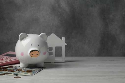 Piggy bank, house model, calculator and money on white wooden table. Space for text