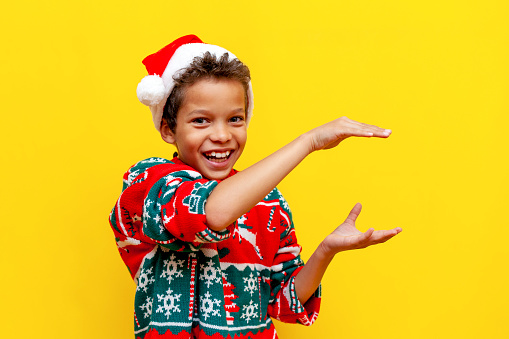 funny african american boy in christmas clothes holds empty hands in his hands on yellow isolated background, 9 year old child in santa hat shows and holds empty hands