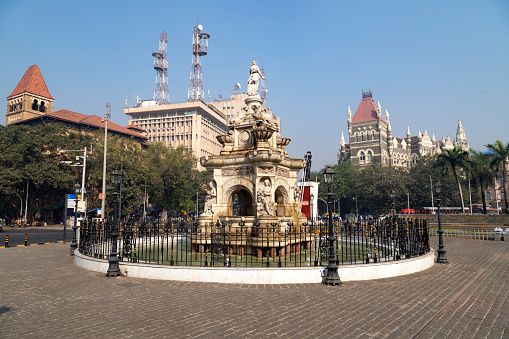 This image captures the iconic Flora Fountain, located at the heart of Mumbai in the Fort business district. Named after the Roman Goddess of Flowers, the fountain is a magnificent stone structure from the colonial era, showcasing intricate carvings and statues. In the photograph, the fountain stands as a striking centerpiece amidst the bustling city life, with its water jets adding a sense of tranquility to the busy surroundings. The image aims to depict the blend of Mumbai’s historical heritage with its modern urban landscape, highlighting Flora Fountain as not just a landmark, but a symbol of the city's enduring charm and a popular meeting point for both locals and tourists.