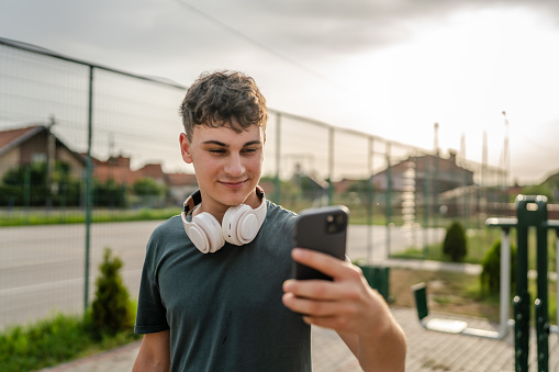 One man caucasian young male stand at outdoor open training park gym use mobile phone smartphone with headphones send messages texting or browse internet online app for training real person copy space