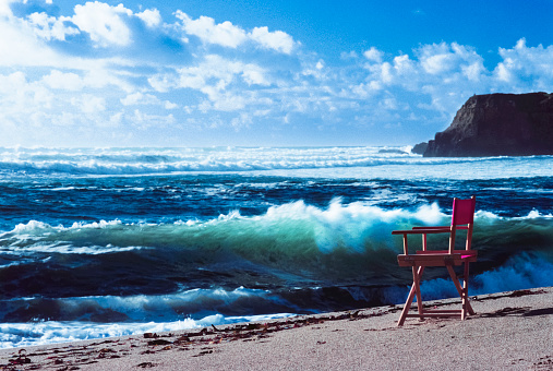 Medium shot of vacant red chair on an ocean beach, overlooking large ocean waves.\n\nTaken on Davenport Beach, Davenport, California, USA