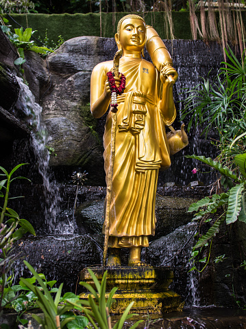 In Bangkok, Thailand, a serene golden Buddha statue stands gracefully amid rocks, embraced by a gentle drizzle outside a temple. This tranquil scene captures the statue's presence amidst the natural elements in this spiritual setting.