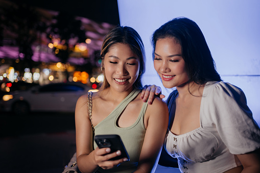 Two smiling friends are looking at the phone in the city at night.