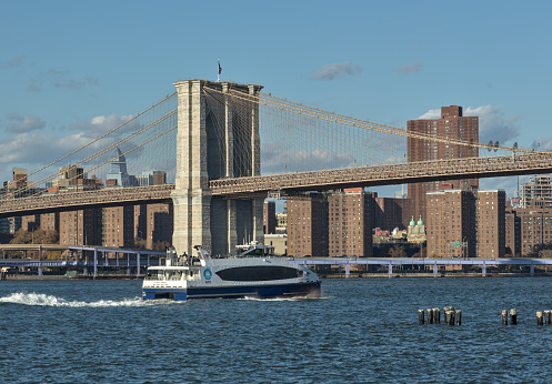 Brooklyn, NY - Nov 19, 2023: NYC ferry passes in front of the Brooklyn Bridge, a famous NYC travel landmark on the Hudson River in Dumbo, New York City.