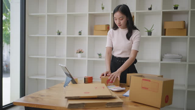 Young Asian woman takine online order through her computer tablet while packing her products in packing boxes