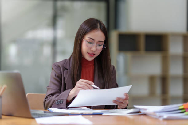 istock Young Asian businesswoman using laptop computer, taking notes, working project in modern office. Financier working with documents at workplace. 1803452575
