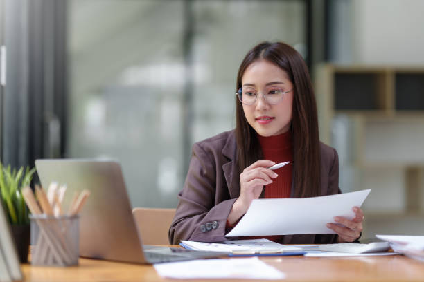 istock Young Asian businesswoman using laptop computer, taking notes, working project in modern office. Financier working with documents at workplace. 1803452589