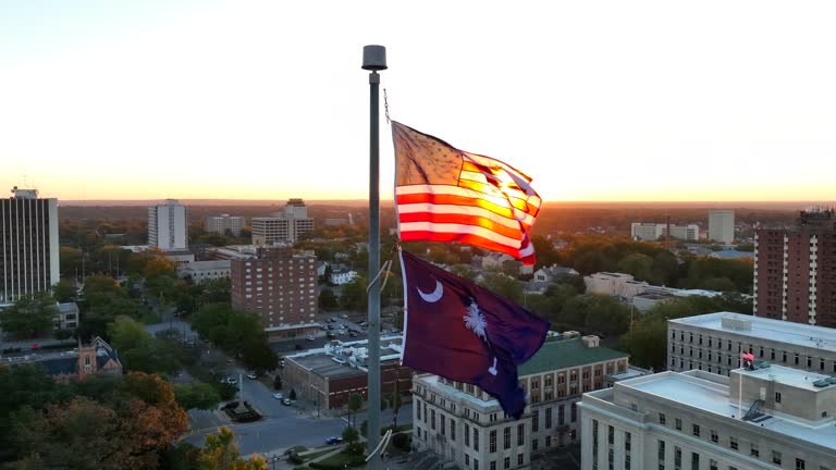 American and South Carolina flags waving atop capitol building in downtown Columbia, SC. Aerial orbit shot with government buildings during bright sunrise.