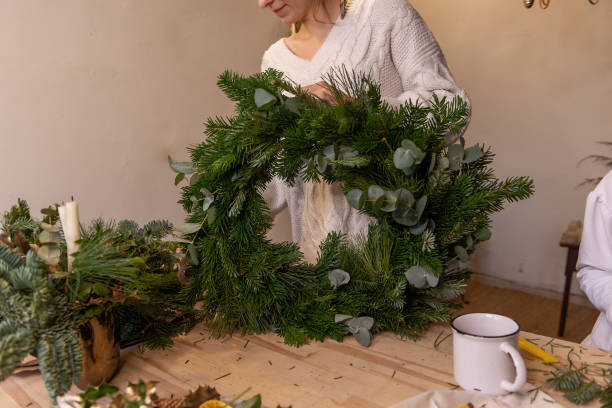 Faceless young woman in knitted sweater makes fluffy green Christmas wreath from natural spruce pine Faceless young woman in knitted sweater makes fluffy, green Christmas wreath from natural spruce pine, eucalyptus branches. Warm, winter atmosphere in rustic interior. Authentic garland of light bulbs personalised ornament wreath stock pictures, royalty-free photos & images