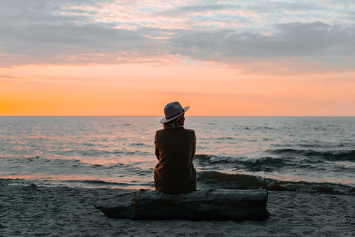 Lonely adult woman wearing a coat and hat sitting alone on a log by the sea at sunset looking away, back view.