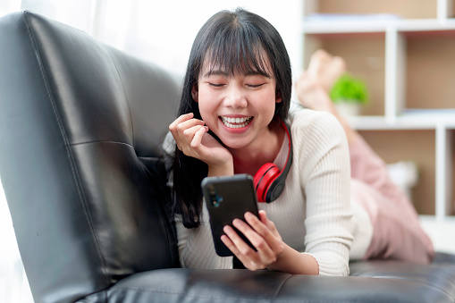 Cute beautiful young Asian woman lying on the sofa relaxing on the sofa playing social media wearing headphones listening to music happily at home.