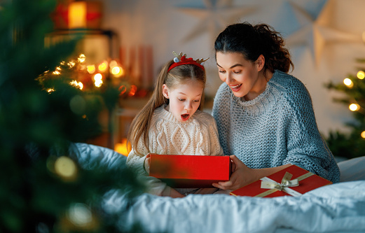 Merry Christmas and Happy Holidays. Cheerful mom and her cute daughter girl exchanging gifts. Parent and little child having fun near tree indoors. Loving family with presents in room.